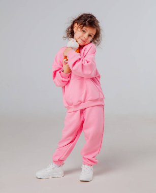 Genç bir çocuk kendine güvenerek şık pembe bir salon kıyafeti içinde duruyor, renkli doldurulmuş bir hayvanı tutuyor. Sahne parlak, minimalist bir stüdyo arka planı içeriyor..