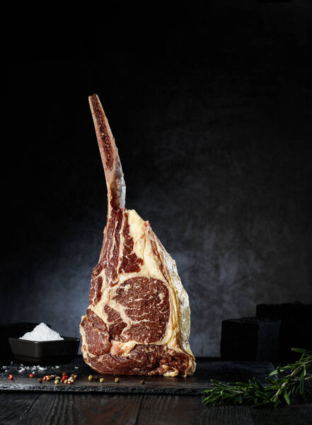 A large tomahawk steak stands upright on a dark stone surface, highlighted by its impressive marbling. Nearby, a small bowl holds salt, while fresh herbs add a touch of color.
