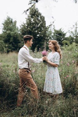 Tarz sahibi genç bir çift, kızın elini tutan, gülümseyen ve birbirine bakan bir erkek.