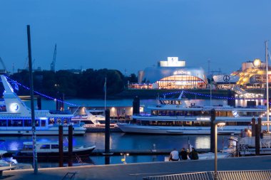 HAMBURG, Almanya - 15 AĞUSTOS 2024 Hamburg Limanı 'nda akşam. Ön planda aydınlatılmış gemiler, arka planda Kar Kraliçesi müzikalinin sahnelendiği tiyatrolar var.