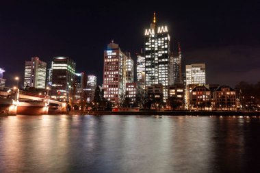 FRANKFURT (MAIN), GERMANY - DECEMBER 3, 2024: A night view of the Frankfurt am Main skyline, featuring the Main River reflecting the lights of the modern skyscrapers in the Bankenviertel and a bridge in the foreground clipart