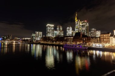 FRANKFURT (MAIN), GERMANY - 3 Aralık 2024: Frankfurt am Main siluetinin gece panoramik görüntüsü, Bankenviertel 'deki modern gökdelenlerin parlak ışıklarını ve nehir kıyısı boyunca uzanan tarihi binaları yansıtıyor.