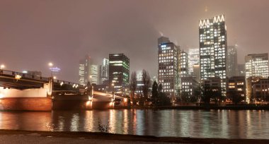 FRANKFURT, Almanya - 2 ARALIK. 2024: Geceleri aydınlık gökdelenleri, köprüsü ve nehri olan modern şehir manzarası