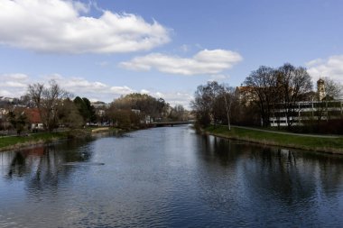 Almanya, Sigmaringen 'den akan Tuna Nehri' nin berrak, parçalı bulutlu gökyüzünün altında sakin bir manzarası.
