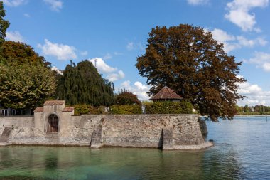Almanya 'nın Konstanz kentindeki Constance Gölü' nün (Bodensee) sakin kıyı şeridinin parlak mavi gökyüzünün altında pofuduk beyaz bulutlarla kaplı bir görüntüsü. Ön planda, küçük ve dayanıklı taş bir duvar var.