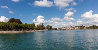 Konstanz, Almanya 'daki sakin Constance Gölü' nün (Bodensee) geniş panoramik manzarası, dağınık beyaz bulutlarla kaplı parlak mavi bir gökyüzünün altında. Sakin, turkuaz sular ön plana doğru uzanıyor.