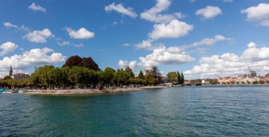 Konstanz, Almanya 'daki sakin Constance Gölü' nün (Bodensee) geniş panoramik manzarası, dağınık beyaz bulutlarla kaplı parlak mavi bir gökyüzünün altında. Sakin, turkuaz sular ön plana doğru uzanıyor.