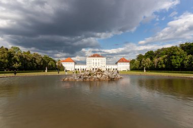 MÜZİK, GERMANY - 17 MAYIS 2025 Münih, Almanya 'daki ikonik Nymphenburg Sarayı' nın resimli geniş açılı manzarası dramatik bir gökyüzü altında sakin saray gölü boyunca görüldü.