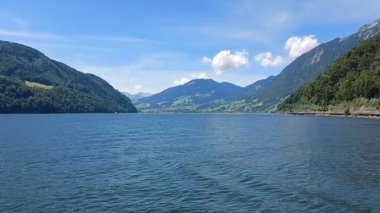 İsviçre 'deki Lucerne Gölü' nün (Vierwaldstttersee) göz kamaştırıcı panoramik manzarası, canlı, mavi bir gökyüzü altında, tüylü beyaz bulutlarla kaplı.