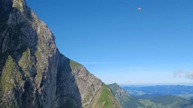 İsviçre Alpleri 'ndeki Pilatus Dağı' ndaki bir teleferiğin parlak mavi gökyüzünün altından nefes kesici yüksek açılı görüntüsü. Yüksek bir dağın heybetli, engebeli kaya yüzeyi çerçevenin sol tarafına hükmediyor.