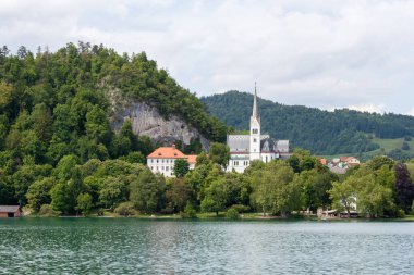 Bled Gölü 'nün sakin suları boyunca manzaralı bir manzara, yemyeşil tepelerin eteklerindeki büyüleyici Bled kasabasını gözler önüne seriyor. Belirgin bir şekilde St. Martin Kilisesi 'nin çan kulesi sergilenmektedir.