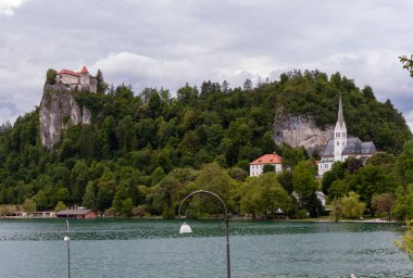 Slovenya 'nın Bled Gölü' nü çevreleyen ikonik simgelerin panoramik bir görüntüsü. Solda, Kanlı Kale yoğun yeşil ormanlarla çevrili dramatik kayalık bir uçurumun üzerinde görkemli bir şekilde tünemektedir. Sağda, St. Martin 'in zarif kilisesi