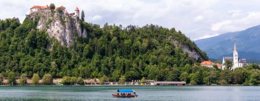 BLED, SLOVENIA - 20 Mayıs 2025: Slovenya 'nın Bled Gölü' nün geniş panoramik manzarası, bulutlu gökyüzü altında ikonik simgelerini barındırıyor: Bled Castle, St. Martin Kilisesi ve geleneksel pletna teknesi