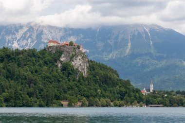 Slovenya 'nın Bled Gölü' nü çevreleyen ikonik simgelerin panoramik bir görüntüsü. Solda, Kanlı Kale yoğun yeşil ormanlarla çevrili dramatik kayalık bir uçurumun üzerinde görkemli bir şekilde tünemektedir. Sağda, St. Martin 'in zarif kilisesi