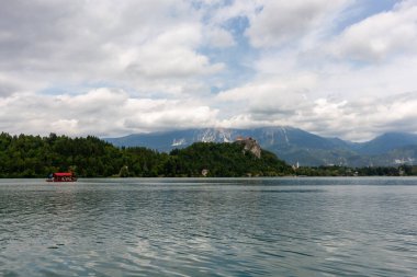 Slovenya 'nın Bled Gölü' nün sakin ve dalgalı sularında beyaz bulutlarla dolu dramatik bir gökyüzünün altında geniş, manzaralı bir manzara. Orta sahada, kırmızı kanopili ve yolcuları olan geleneksel bir pletna tekne.