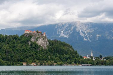 Slovenya 'nın Bled Gölü' nü çevreleyen ikonik simgelerin panoramik bir görüntüsü. Solda, Kanlı Kale yoğun yeşil ormanlarla çevrili dramatik kayalık bir uçurumun üzerinde görkemli bir şekilde tünemektedir. Sağda, St. Martin 'in zarif kilisesi
