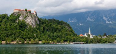 Slovenya 'nın Bled Gölü' nü çevreleyen ikonik simgelerin panoramik bir görüntüsü. Solda, Kanlı Kale yoğun yeşil ormanlarla çevrili dramatik kayalık bir uçurumun üzerinde görkemli bir şekilde tünemektedir. Sağda, St. Martin 'in zarif kilisesi