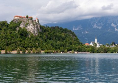 Slovenya 'nın Bled Gölü' nü çevreleyen ikonik simgelerin panoramik bir görüntüsü. Solda, Kanlı Kale yoğun yeşil ormanlarla çevrili dramatik kayalık bir uçurumun üzerinde görkemli bir şekilde tünemektedir. Sağda, St. Martin 'in zarif kilisesi