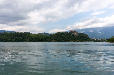 Slovenya 'nın Bled Gölü' nün geniş ve pitoresk bir manzarası, parçalı bulutlu bir gökyüzünün altında. Sakin, dalgalı su ön planı dolduruyor.