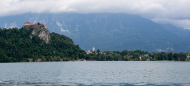 Slovenya, Bled Gölü 'nün geniş panoramik manzarası dramatik ve bulutlu bir gökyüzü altında. Solda, tarihi Kanlı Kale görkemli bir şekilde kayalık bir uçurumun tepesinde, göl kenarına kadar uzanan yemyeşil ormanlarla çevrili.