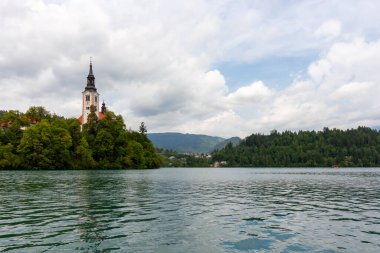 Meryem 'in Varsayımının ikonik kilisesi, belirgin uzun çan kulesiyle, beyaz ve kırmızı cepheleriyle, Bled Adası' nın zümrüt yeşili Bled gölünün sularından yükselen yeşil ağaçların arasında görkemli bir şekilde duruyor.
