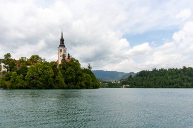 Meryem 'in Varsayımının ikonik kilisesi, belirgin uzun çan kulesiyle, beyaz ve kırmızı cepheleriyle, Bled Adası' nın zümrüt yeşili Bled gölünün sularından yükselen yeşil ağaçların arasında görkemli bir şekilde duruyor.