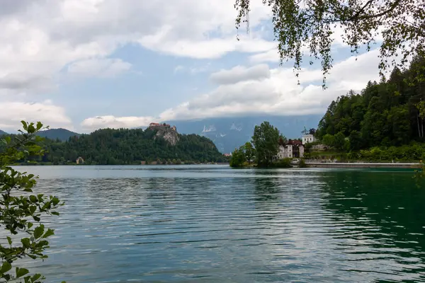 Slovenya 'nın Bled Gölü' nün geniş ve pitoresk bir manzarası, parçalı bulutlu bir gökyüzünün altında. Sakin, dalgalı su ön planı dolduruyor.