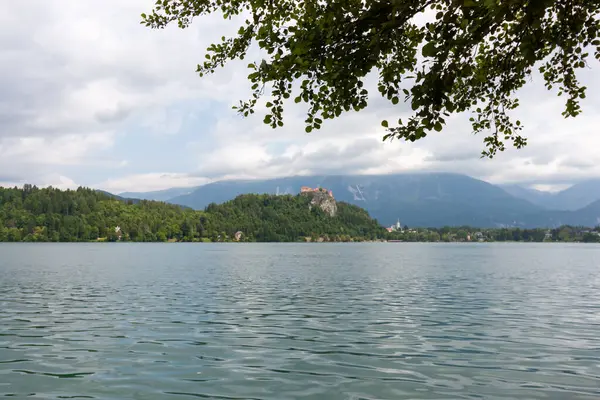 Slovenya 'nın Bled Gölü' nün geniş ve pitoresk bir manzarası, parçalı bulutlu bir gökyüzünün altında. Sakin, dalgalı su ön planı dolduruyor.