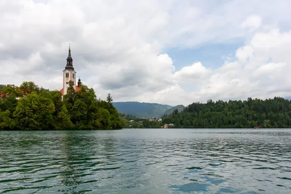 Meryem 'in Varsayımının ikonik kilisesi, belirgin uzun çan kulesiyle, beyaz ve kırmızı cepheleriyle, Bled Adası' nın zümrüt yeşili Bled gölünün sularından yükselen yeşil ağaçların arasında görkemli bir şekilde duruyor.