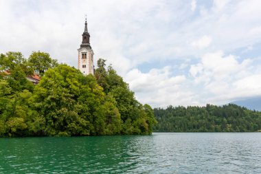Meryem 'in Varsayımının ikonik kilisesi, belirgin uzun çan kulesiyle, beyaz ve kırmızı cepheleriyle, Bled Adası' nın zümrüt yeşili Bled gölünün sularından yükselen yeşil ağaçların arasında görkemli bir şekilde duruyor.