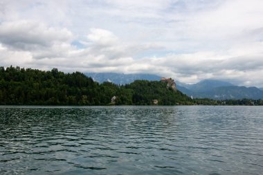 Bled Gölü 'nün pitoresk manzarası ön plandaki yemyeşil yapraklar tarafından çerçevelenmiş ve turkuaz sulara kısa bir bakış sunuluyor. Slovenya