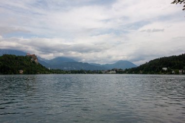 Bled Gölü 'nün pitoresk manzarası ön plandaki yemyeşil yapraklar tarafından çerçevelenmiş ve turkuaz sulara kısa bir bakış sunuluyor. Slovenya