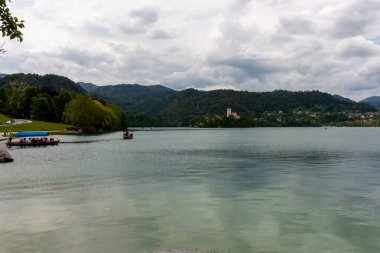 Bled Gölü 'nün pitoresk manzarası ön plandaki yemyeşil yapraklar tarafından çerçevelenmiş ve turkuaz sulara kısa bir bakış sunuluyor. Slovenya