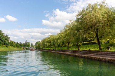 Slovenya 'nın Ljubljana kentindeki Ljubljanica Nehri boyunca sakin ve yeşil bir manzara, aydınlık ve parçalı bulutlu bir günde