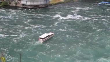 İnsanlarla dolu bir turist teknesi Ren Şelalesi yakınlarındaki çalkantılı, turkuaz sularda yol alır.