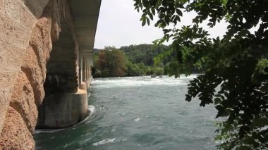 Çalkantılı Ren nehrinin beyaz akıntılı görüntüsünü gösteren bir video çerçevesi, taş bir kemer köprüsünün altından görüldüğü gibi.