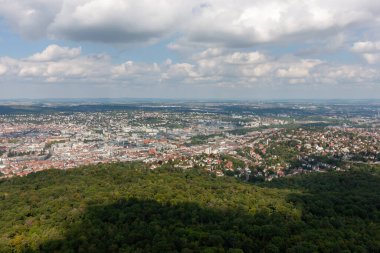 Pofuduk bulutların olduğu güneşli bir günde Stuttgart şehri ve yeşil ormanın havadan görünüşü. Şehir manzarası berrak bir gökyüzünün altına yayılarak şehir ve doğal manzaraların güzel bir karışımını vurguluyor.