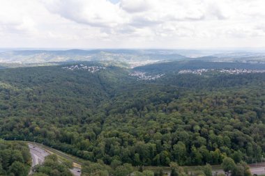 Stuttgart şehrinin yerleşim bölgesini çevreleyen dolambaçlı bir yolu olan yemyeşil bir ormanın havadan görünüşü. Sahne, bulutlu bir gökyüzü altında doğanın ve kentsel yaşamın kesişimini yakalıyor.
