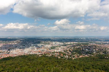 Pofuduk bulutların olduğu güneşli bir günde Stuttgart şehri ve yeşil ormanın havadan görünüşü. Şehir manzarası berrak bir gökyüzünün altına yayılarak şehir ve doğal manzaraların güzel bir karışımını vurguluyor.