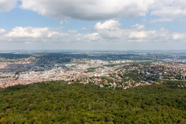 Pofuduk bulutların olduğu güneşli bir günde Stuttgart şehri ve yeşil ormanın havadan görünüşü. Şehir manzarası berrak bir gökyüzünün altına yayılarak şehir ve doğal manzaraların güzel bir karışımını vurguluyor.