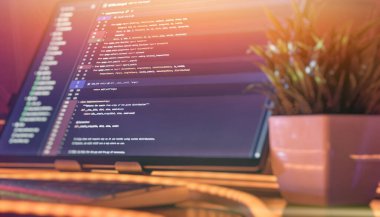 Python kodlu temiz ve organize müteahhit masası, sıcak ortam aydınlatması, RGB klavyesi ve monitörün yanında dekoratif bitki..