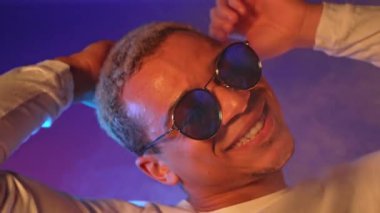 Enerjik Afrikalı Amerikalı adam, neon ışığın altında menekşe arka planda dans ediyor. Beyaz kıvırcık saç stili ve yuvarlak aynalı gözlüklü şık bir adamın portresi. Yüksek kalite 4k görüntü