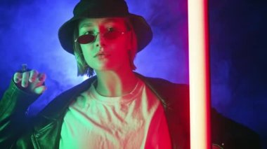 Neon renkli ışıkta dans eden zarif rock kadını. Dumanlı arka plan, gece partisi, disko kulübü. Z jenerasyonu esrarengiz şapkalı güzel bayan, gözlüklü. Yüksek kalite 4k görüntü