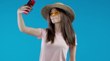 Modaya uygun genç kız mavi arka planda akıllı telefondan selfie çekiyor. Hasır şapkalı ve güneş gözlüklü kadın. Teknoloji, mobil cihaz, sosyal ağ konsepti. Yüksek kalite 4k görüntü