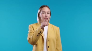 Gülümseyen kadın parmaklarını dudaklarında tutuyor, mavi stüdyo arka planı. Şşş, sır, sessizlik, komplo, dedikodu konsepti olan bir iş kadını. Yüksek kalite 4k görüntü