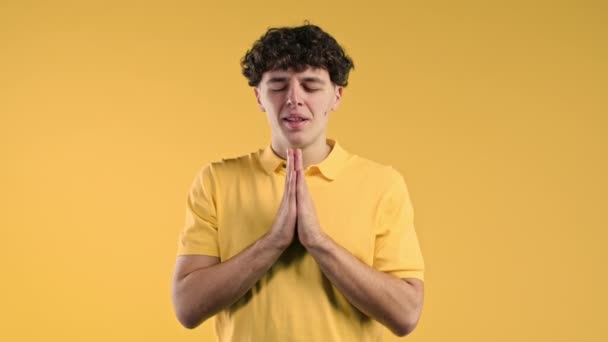 若い男は祈り 彼女を謝罪し始め 彼は有罪です 黄色の背景 誰かに彼の欲望を満たし 助けを求める希望的な祈りの巻き毛の男 高品質4K映像 — ストック動画