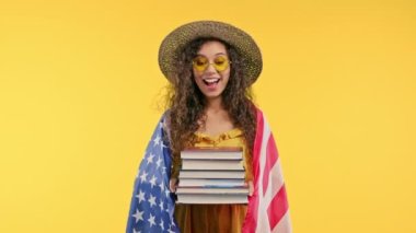 Amerikalı kadın öğrenci, üniversite kütüphanesinden gelen bir yığın kitabı sarı arka planda tutuyor. Mutlu kız gülümser, Amerika 'dan mezun olduğu için mutludur, yurt dışında eğitim kavramı. Yüksek kalite 4k görüntü