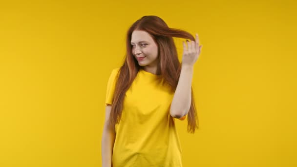 遊び心のある生姜の女性は 黄色のスタジオの背景にカメラにいちゃつく笑顔 かわいい流行の女の子は彼女の長い素晴らしい髪を修正します 高品質4K映像 — ストック動画