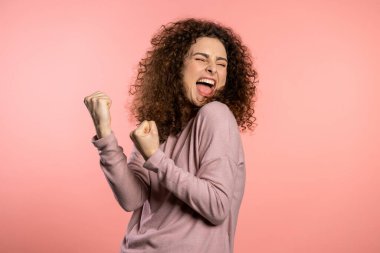 Kıvırcık saçlı ve olağanüstü makyajlı bir kız zaferin evet jestini gösteriyor. Sonuç elde etti, hedeflerine ulaştı. Pembe arka planda heyecanlı mutlu bir kadın..