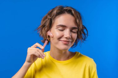 Mavi arka planda gül kuvars taşıyla yüz masajı yapan genç bir kadın. Yüz bakımı, güzellik ritüelleri, kozmetoloji, yaşlanmayı önleme ve kırışıklık önleme tedavisi. Yüksek kalite fotoğraf